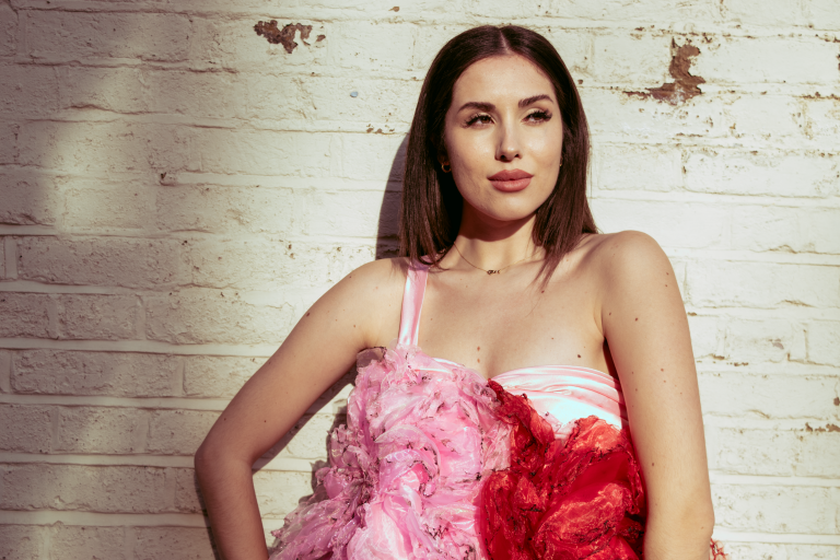 Alba de la Llave in pink and red floral dress, sunlit against a white brick wall.