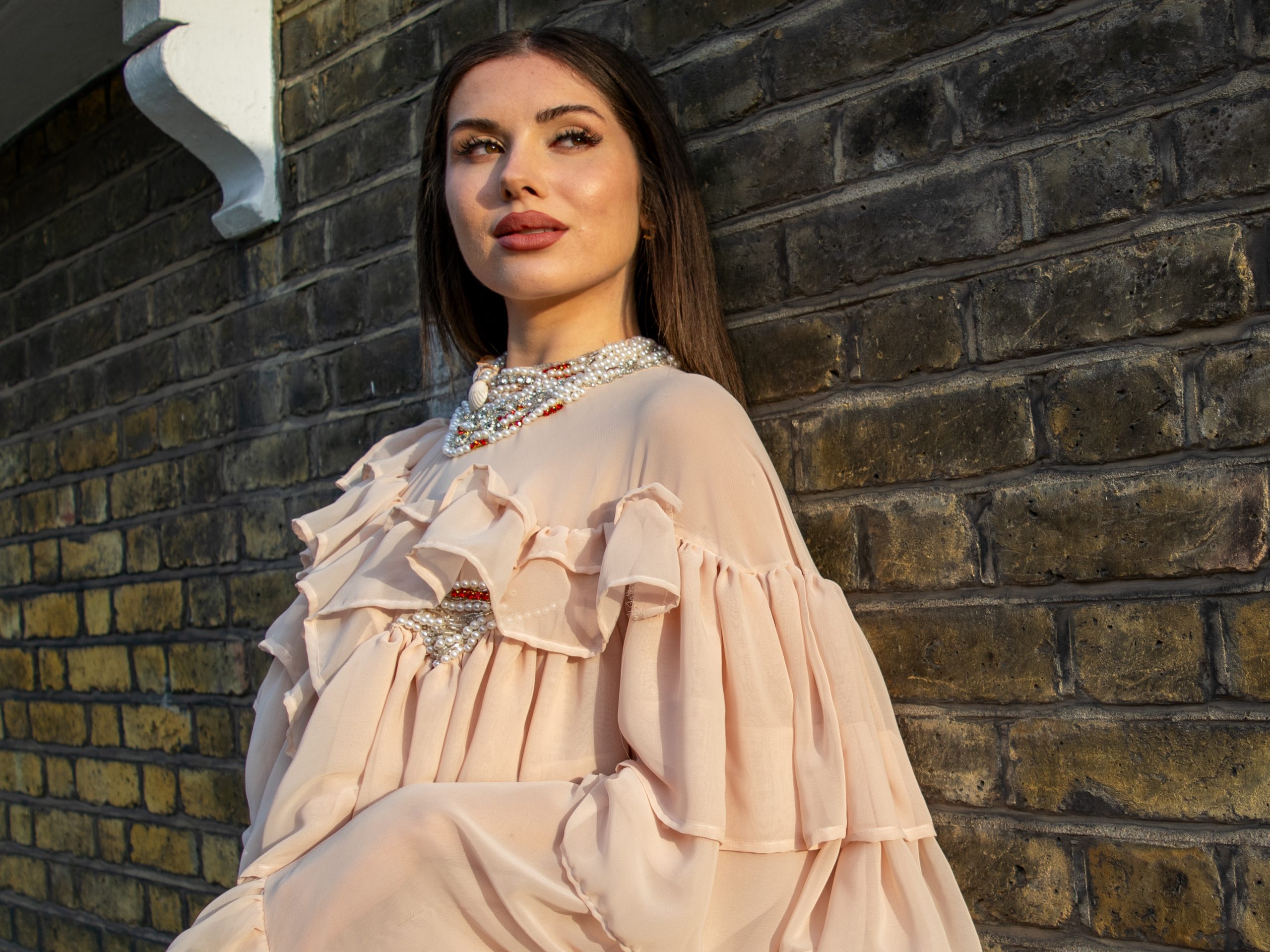 Alba De La Llave stands by a brick wall in golden light, wearing a ruffled couture dress.