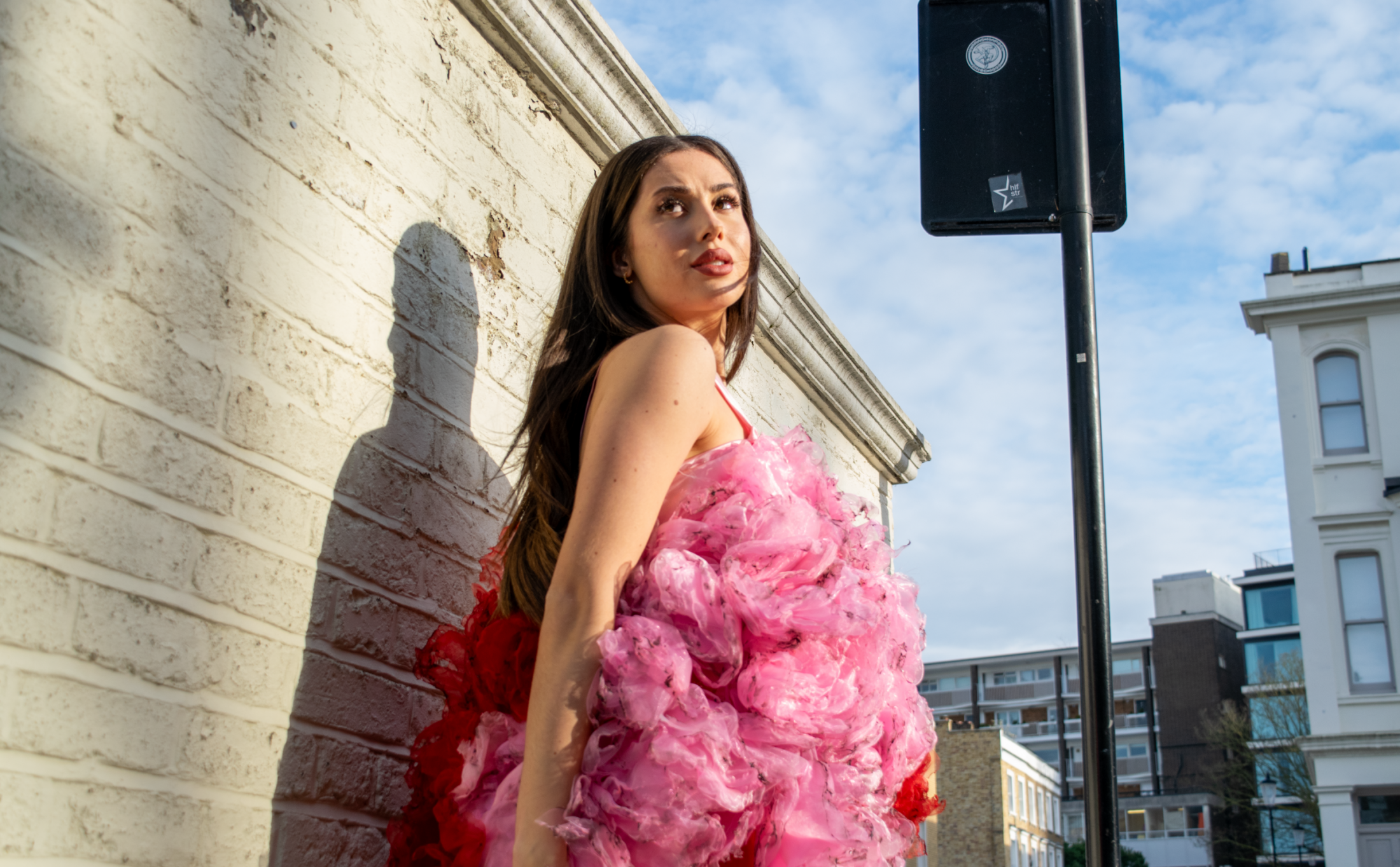 Alba de la Llave stands tall in a floral dress against a white brick wall, street sign above and sunlit London backdrop.